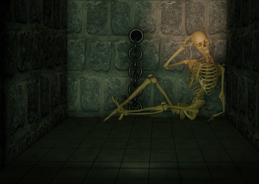 A skeleton shackled to a dungeon wall. Leaned up against the corner and holding his head in a contemplative pose.